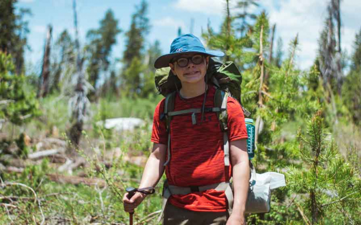 a young person wearing a backpack and hat smiles at the camera amid a wooded area